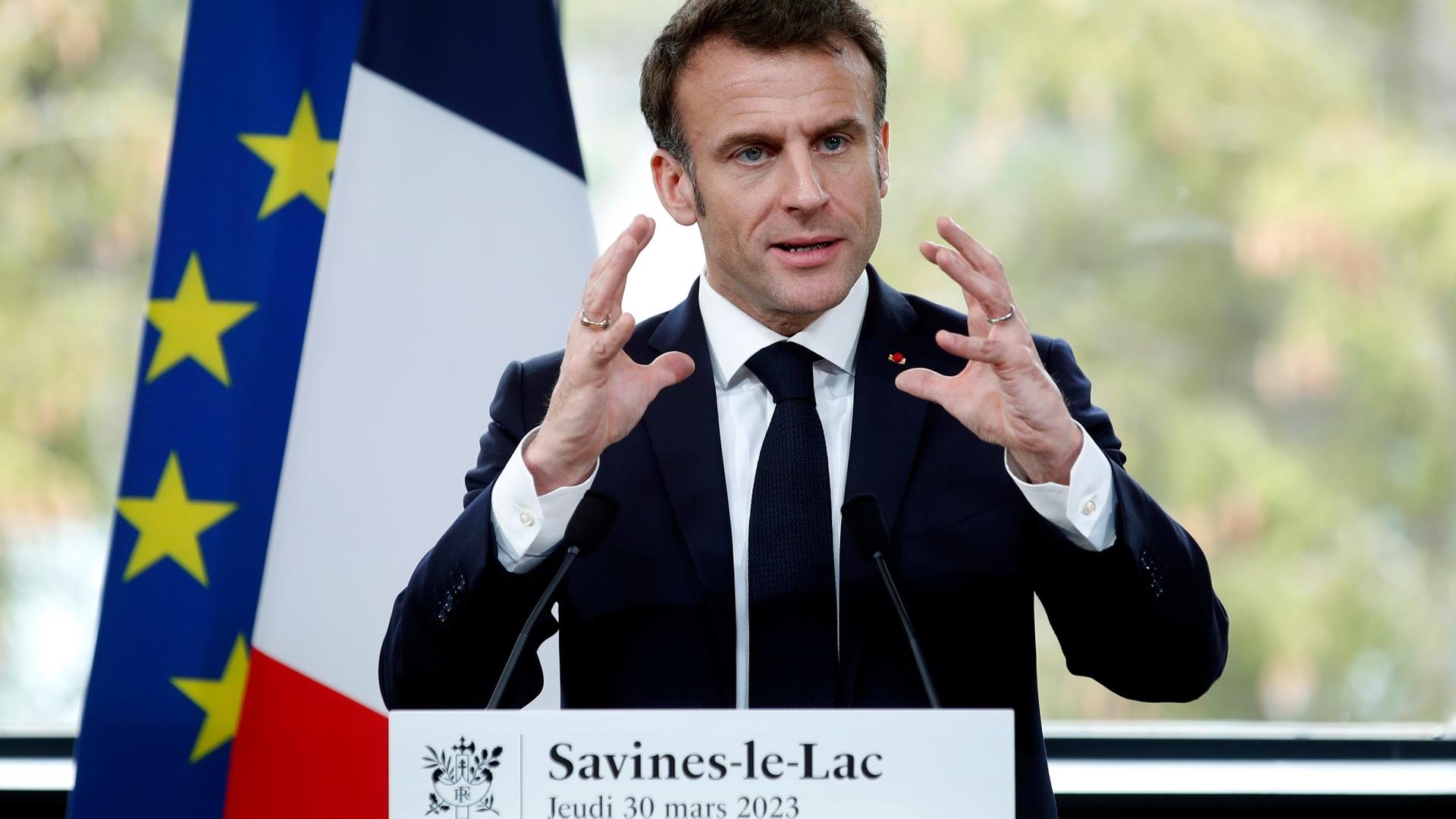 Der französische Präsident Macron steht hinter zwei Mikrofonen und hält gestikulierend eine Rede. Hinter ihm sind eine französische und eine EU-Flagge zu sehen.
