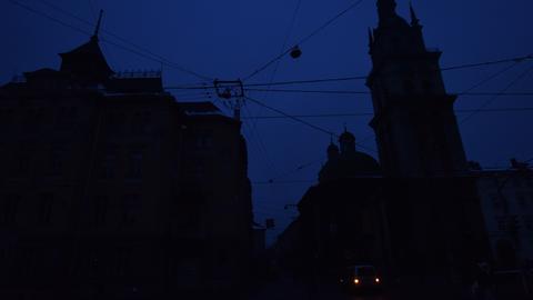 Eine Stadtlandschaft in fast kompletter Dunkelheit. Nur an einer Stelle sind zwei Lichter zu sehen, sonst zeichnen sich lediglich dunkle Gebäudesilhouetten gegen den dunkelblauen Himmel ab.