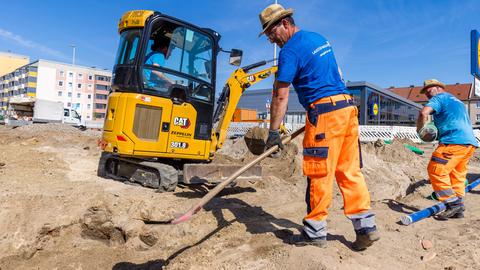 Arbeiter einer Tiefbaufirma arbeiten mit Schaufeln auf einer Baustelle. Im Hintergrund steht ein Mini-Bagger.