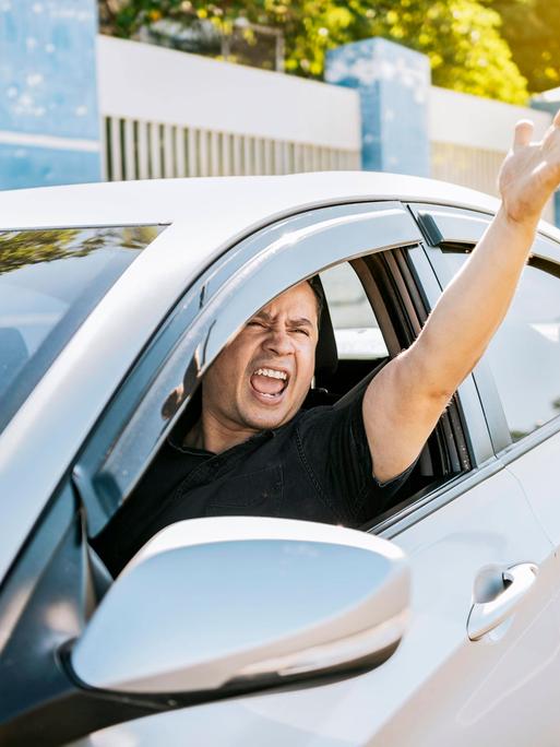 Ein wütender Fahrer schreit einen anderen Verkehrsteilnehmer aus dem Auto heraus an und gestikuliert durch das Fenster heraus. Ein wütender Fahrer schreit einen anderen Verkehrsteilnehmer aus dem Auto heraus an und gestikuliert durch das Fenster heraus.