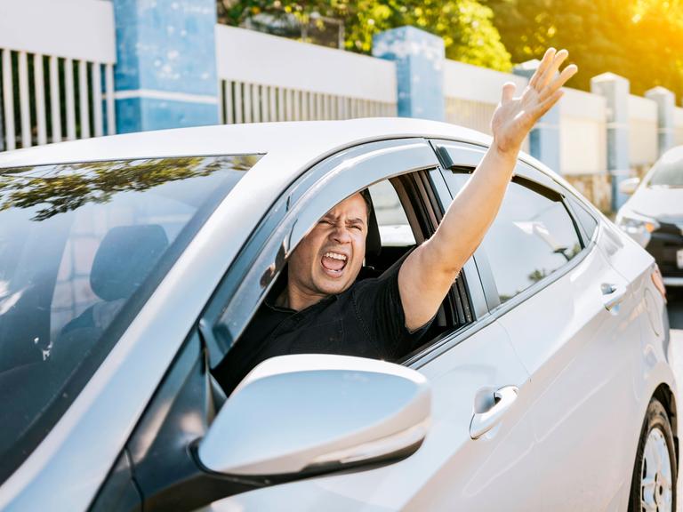 Ein wütender Fahrer schreit einen anderen Verkehrsteilnehmer aus dem Auto heraus an und gestikuliert durch das Fenster heraus. Ein wütender Fahrer schreit einen anderen Verkehrsteilnehmer aus dem Auto heraus an und gestikuliert durch das Fenster heraus.