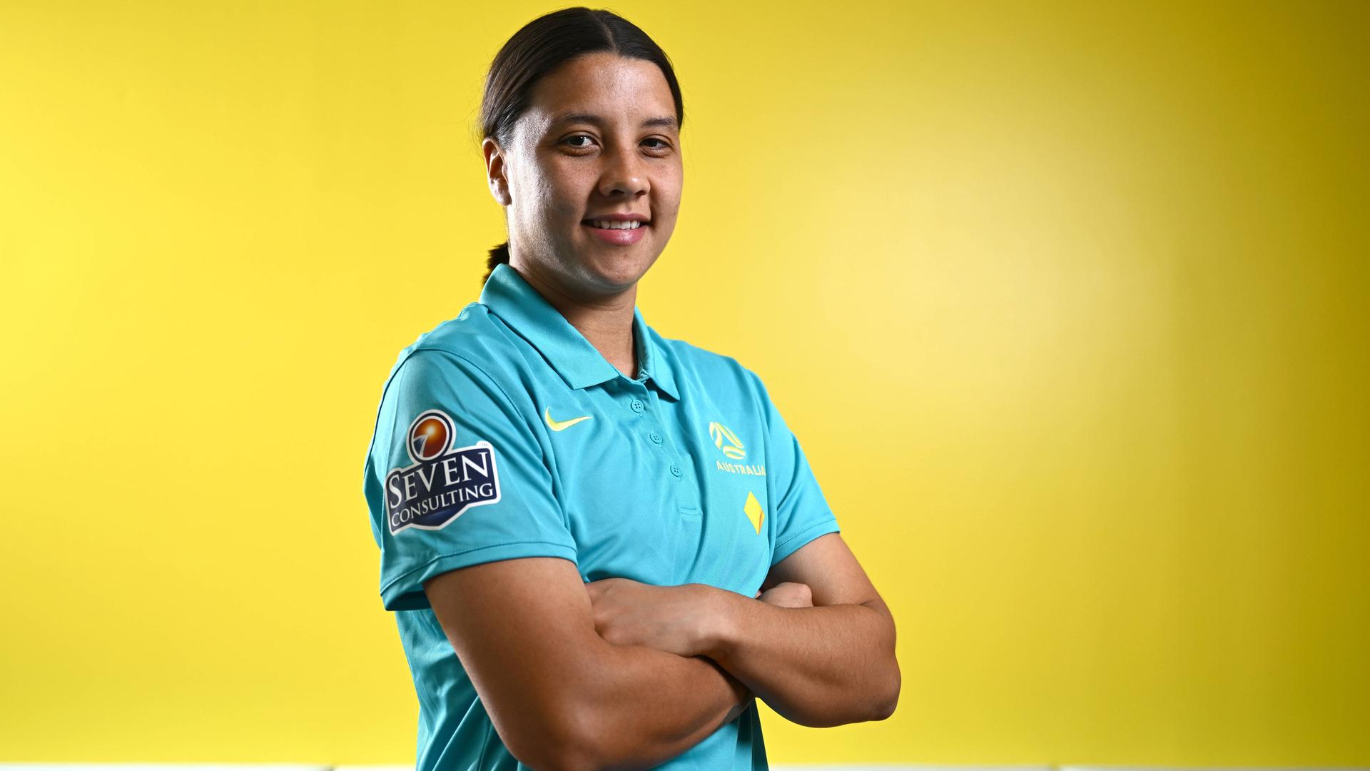 Sam Kerr steht mit verschränkten Armen und siegesgewissem Lächeln vor einer gelben Wand.