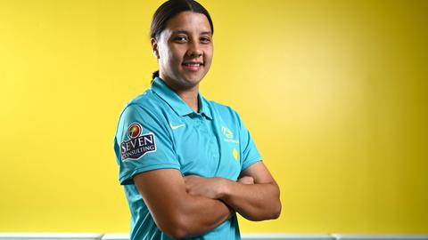 Sam Kerr steht mit verschränkten Armen und siegesgewissem Lächeln vor einer gelben Wand.