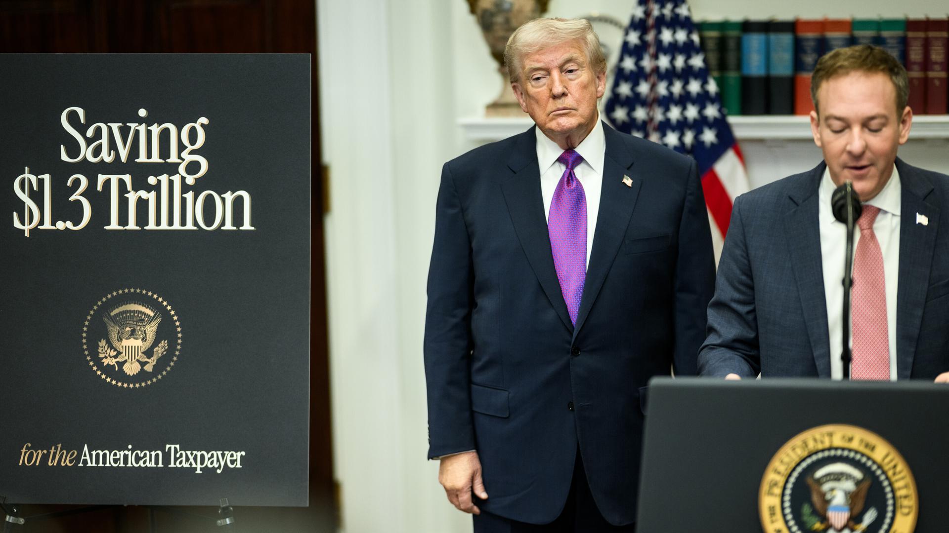Zeldin spricht an einem Pult mit dem Präsidentensiegel; Trump steht daneben und schaut ernst vor suich hin. Links von den beiden eine Tafel mit der Aufschrift "Saving 1,3 Trillion Dollars". 
