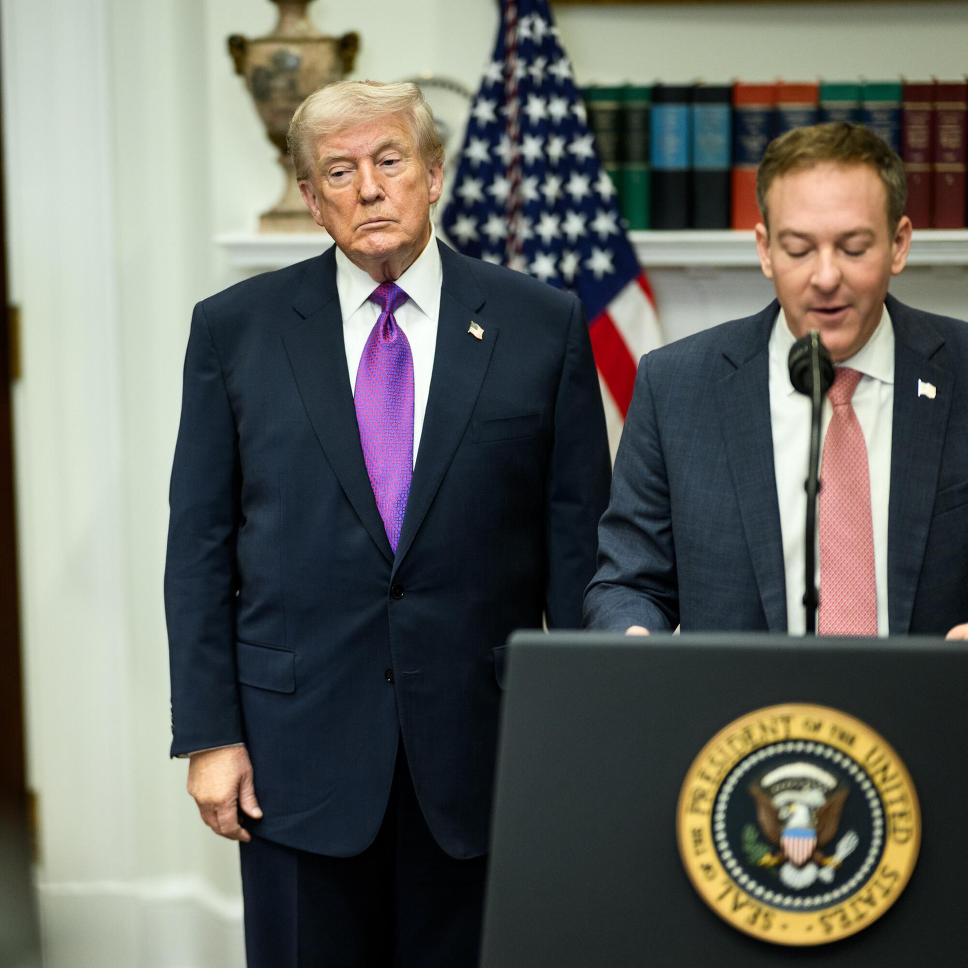 Zeldin spricht an einem Pult mit dem Präsidentensiegel; Trump steht daneben und schaut ernst vor suich hin. Links von den beiden eine Tafel mit der Aufschrift "Saving 1,3 Trillion Dollars".  