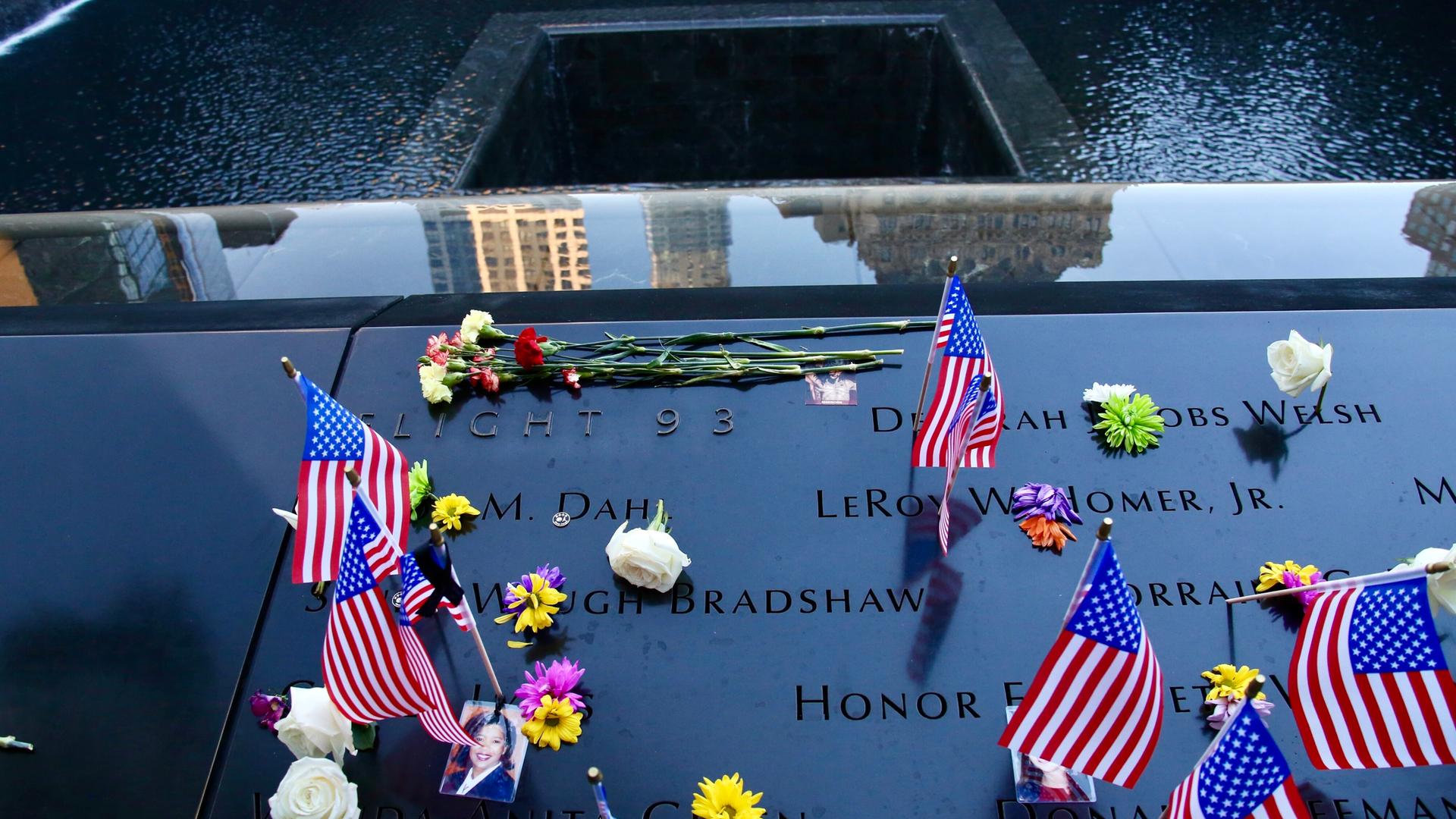 Das Bild zeigt eine Gedenktafel mit Namen der zahlreichen Opfer. Es liegen außerdem Blumen und US-amerikanische Flaggen darauf.