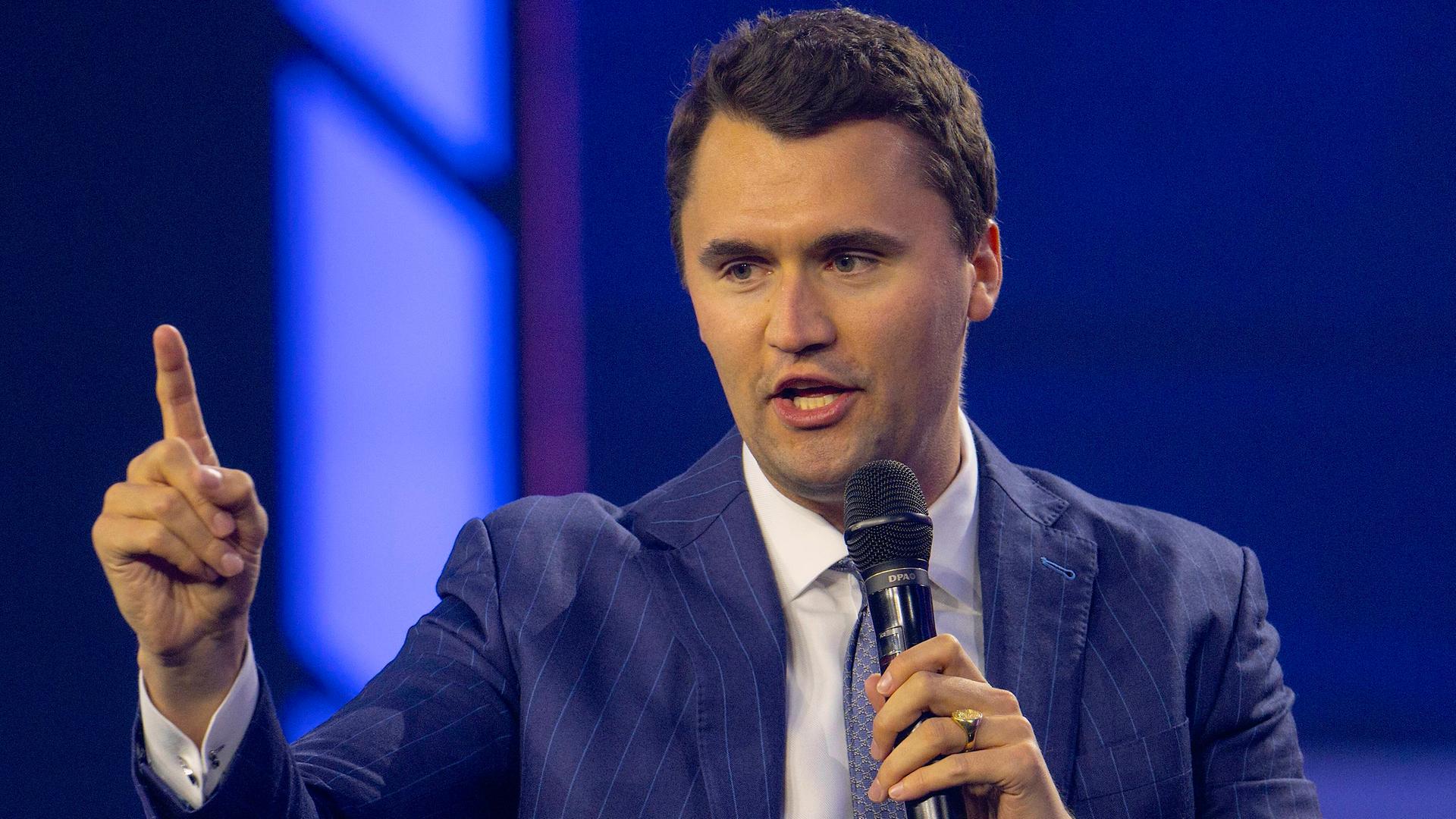 Charlie Kirk im blauen Anzug mit erhobenem Zeigefinger, während er in ein Mikrofon spricht.