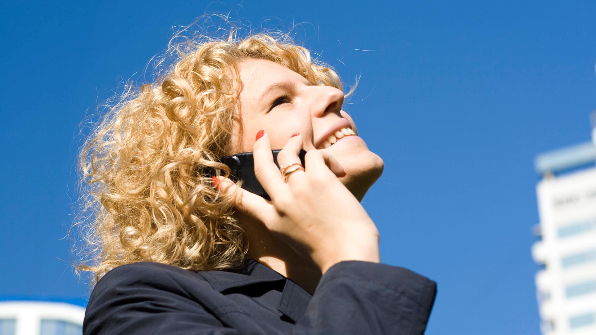 Frau mit blonden Locken vor einem tiefblauen Himmel telefoniert mit einem Handy am Ohr mit glücklichem Lächeln. Frau mit blonden Locken vor einem tiefblauen Himmel telefoniert mit einem Handy am Ohr mit glücklichem Lächeln.