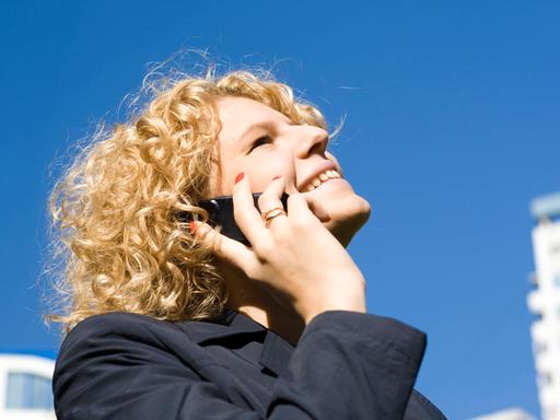 Frau mit blonden Locken vor einem tiefblauen Himmel telefoniert mit einem Handy am Ohr mit glücklichem Lächeln.