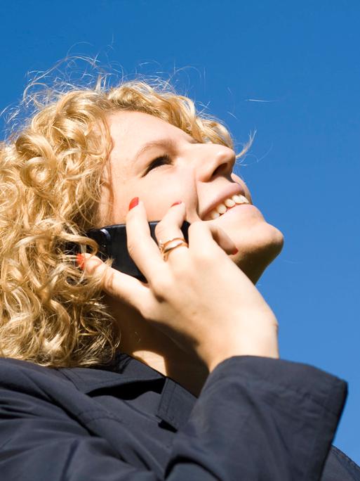 Frau mit blonden Locken vor einem tiefblauen Himmel telefoniert mit einem Handy am Ohr mit glücklichem Lächeln.