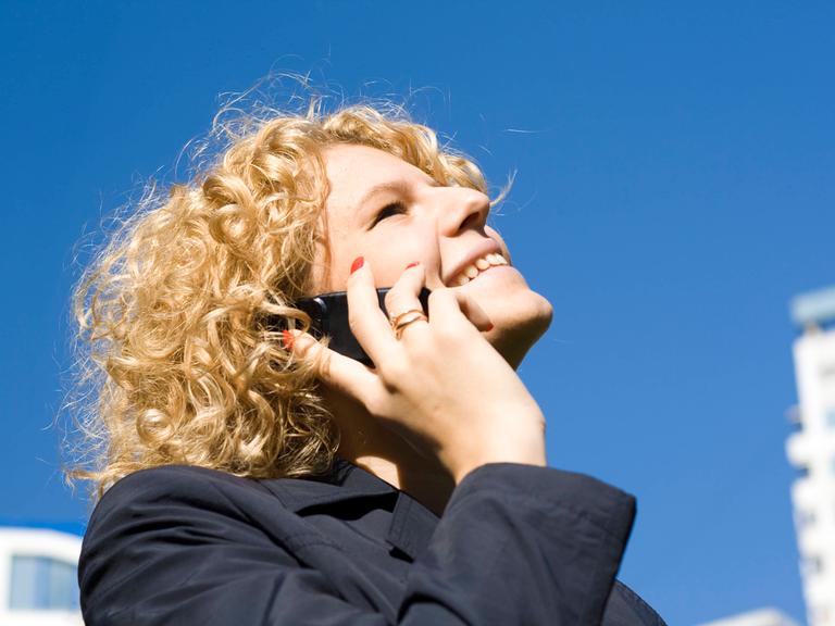 Frau mit blonden Locken vor einem tiefblauen Himmel telefoniert mit einem Handy am Ohr mit glücklichem Lächeln.