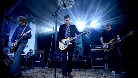 Thorsten Nagelschmidt steht während eines Auftritts mit seiner Band Muff Potter auf der Bühne. Er spielt Gitarre und trägt ein Hemd mit Blumenmuster zum Anzug.