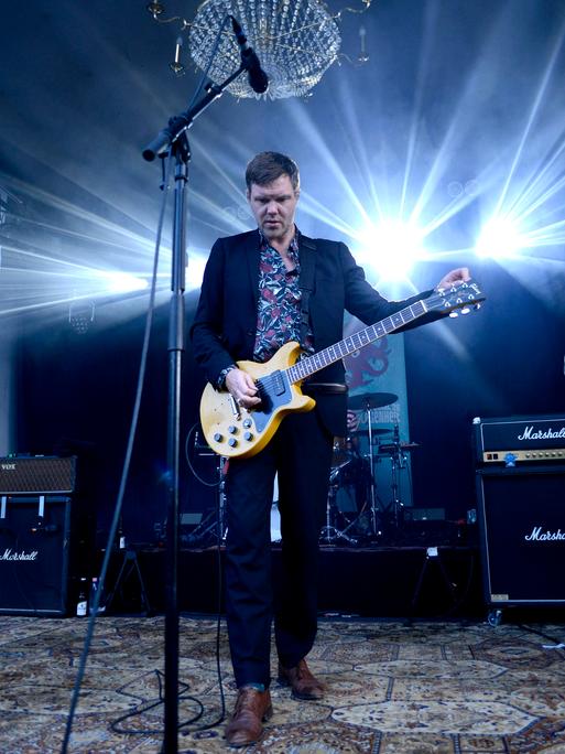 Thorsten Nagelschmidt steht während eines Auftritts mit seiner Band Muff Potter auf der Bühne. Er spielt Gitarre und trägt ein Hemd mit Blumenmuster zum Anzug.