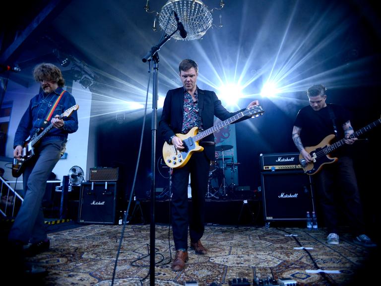 Thorsten Nagelschmidt steht während eines Auftritts mit seiner Band Muff Potter auf der Bühne. Er spielt Gitarre und trägt ein Hemd mit Blumenmuster zum Anzug.