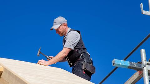 Ein Zimmermann sitzt auf einem Dach und schlägt mit dem Hammer einen Nagel ein.