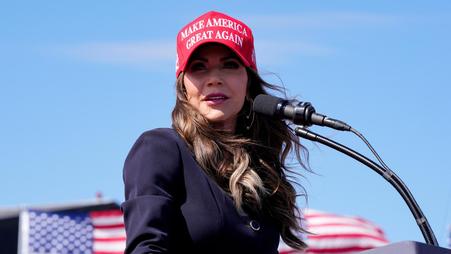 Kristi Noem hält eine Rede unter freiem Himmel. Sie trägt die gleiche rote Kappe wie Donald Trump. Darauf steht "Make America great again".