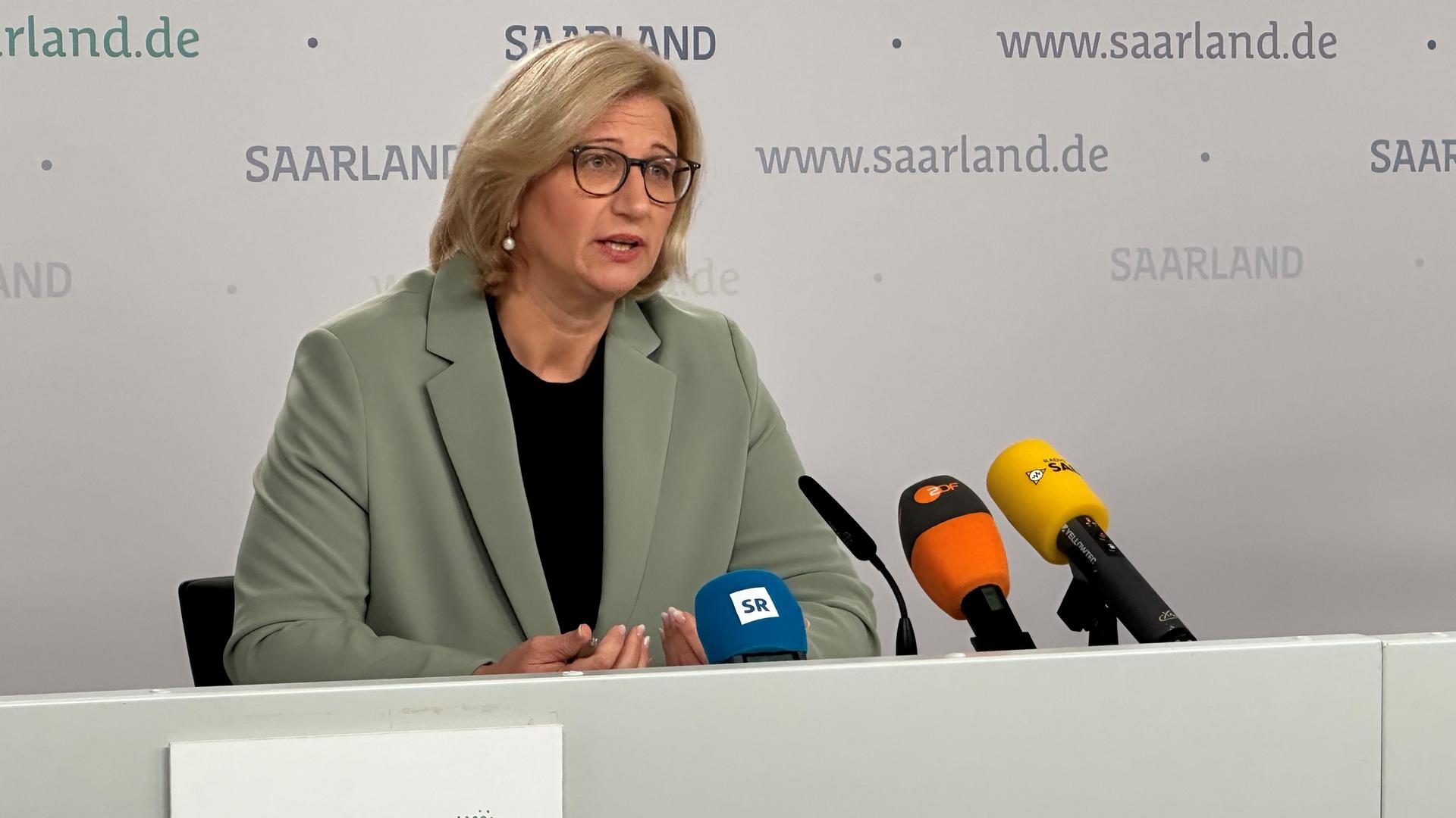 Rehlinger sitzt an einem Tisch und spricht in mehrere Mikrofone. Dahinter eine graue Wand mit Schriftzügen des Saarlands.