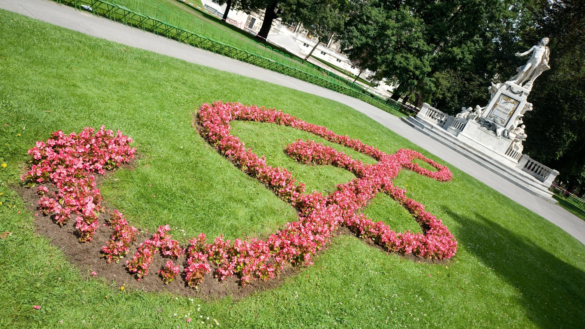 Auf dem Rasen vor dem weißen Mozartdenkmal in Wien sind rosafarbene Blumen angepflanzt, die einen überdimensionalen Notenschlüssel ergeben.