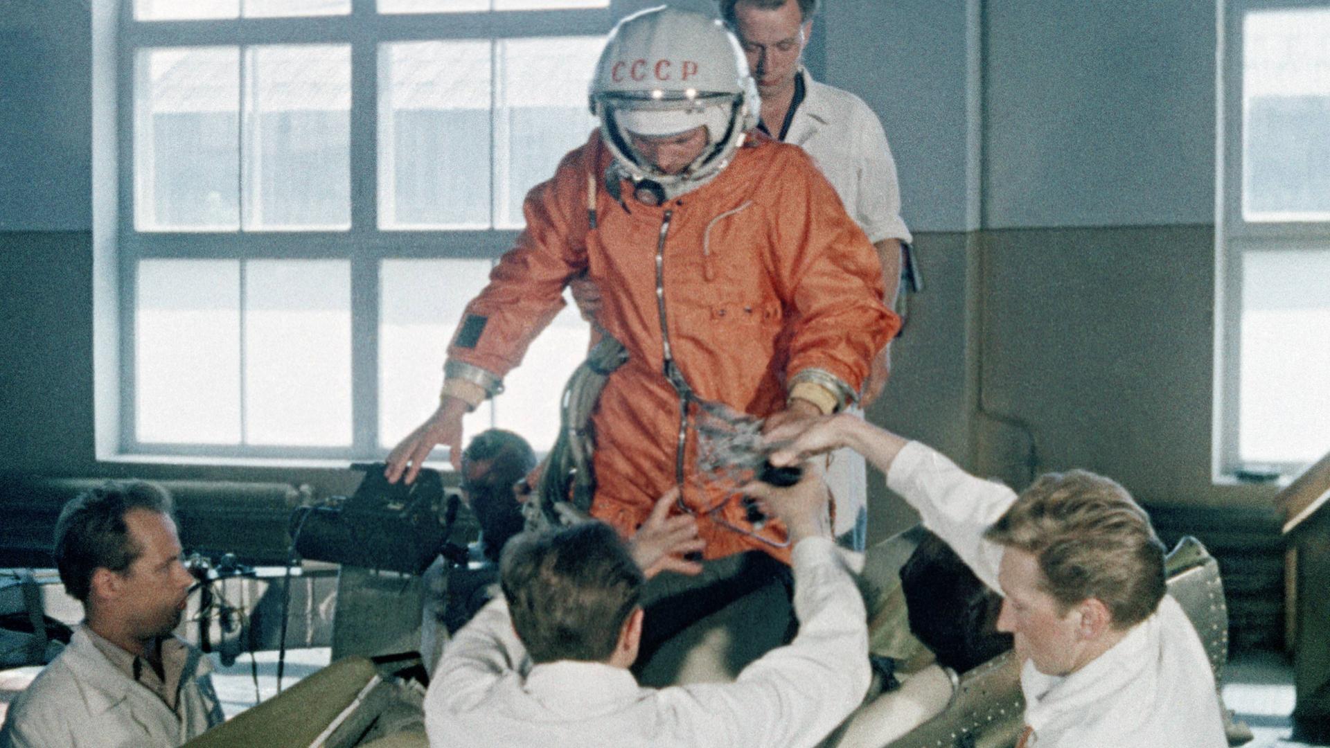 Vorbereitung des ersten Kosmonauten Juri Gagarin auf den Raumflug. Er trägt einen orangenen Anzug und ist umgeben von Männern in weißen Kitteln, die ihn zu stützen scheinen. Vorbereitung des ersten Kosmonauten Juri Gagarin auf den Raumflug. Er trägt einen orangenen Anzug und ist umgeben von Männern in weißen Kitteln, die ihn zu stützen scheinen.