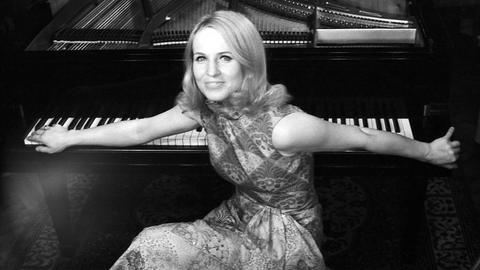 Schwarzweiß Bild der deutschen Pianistin und Hochschullehrerin Annerose Schmidt. Sie sitzt auf einem Hocker am Klavier und hat den Rücken zu dem Instrument gewandt und ihre Arme auf den Tasten ausgebreitet. 