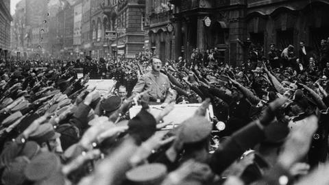 Adolf Hitler fährt stehend auf einem Wagen. An beiden Straßenseiten jubeln ihm Menschen zu. Sie zeigen den Hitlergruß.