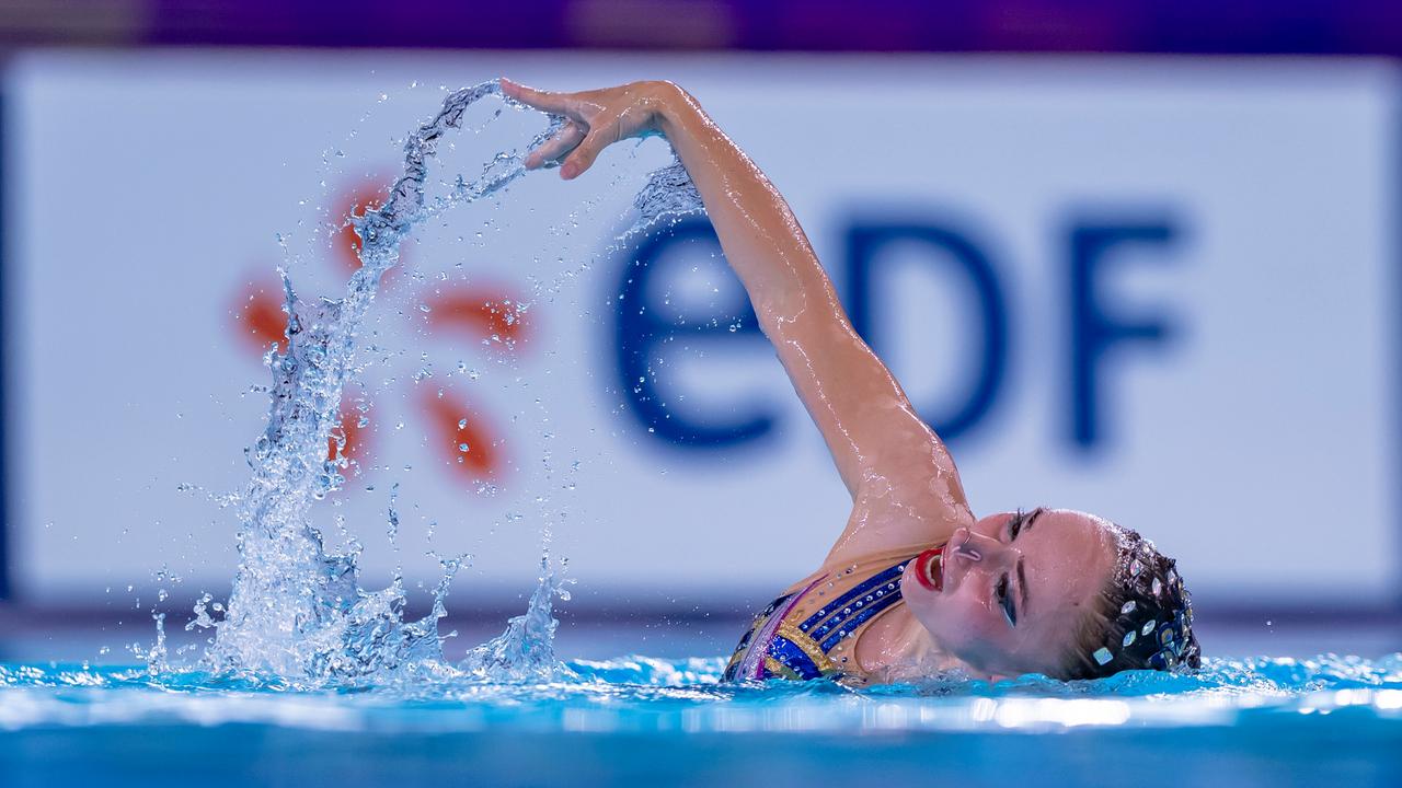Snychronschwimmerin Klara Bleyer hebt bei der Freien Kür im Wasser einen Arm, im Hintergrund das Logo eines Sponsors.