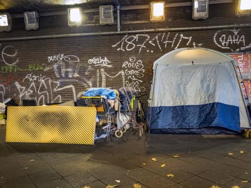 Unter einer Brücke am Stuttgarter Platz in Berlin, ist ein grau-blaues Zelt aufgebaut. Ein Einkaufswagen mit Decken steht daneben. Eine schmutzige gelbe Matratze lehnt am Einkaufswagen. 14.11.2023