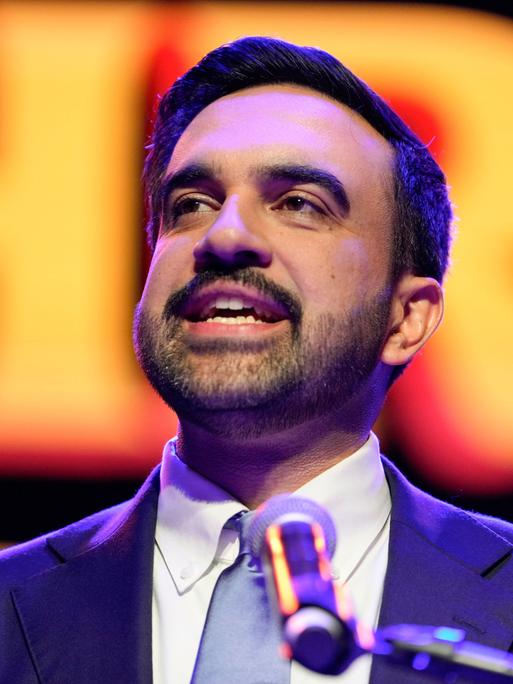 Zohran Mamdani spricht nach seinem Wahlsieg in einem blauen Anzug in New York auf der Bühne in ein Mikrofon. Sein Gesicht ist vor einem orange-roten Hintergrund zu sehen.
