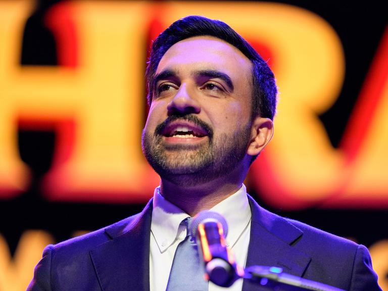 Zohran Mamdani spricht nach seinem Wahlsieg in einem blauen Anzug in New York auf der Bühne in ein Mikrofon. Sein Gesicht ist vor einem orange-roten Hintergrund zu sehen.
