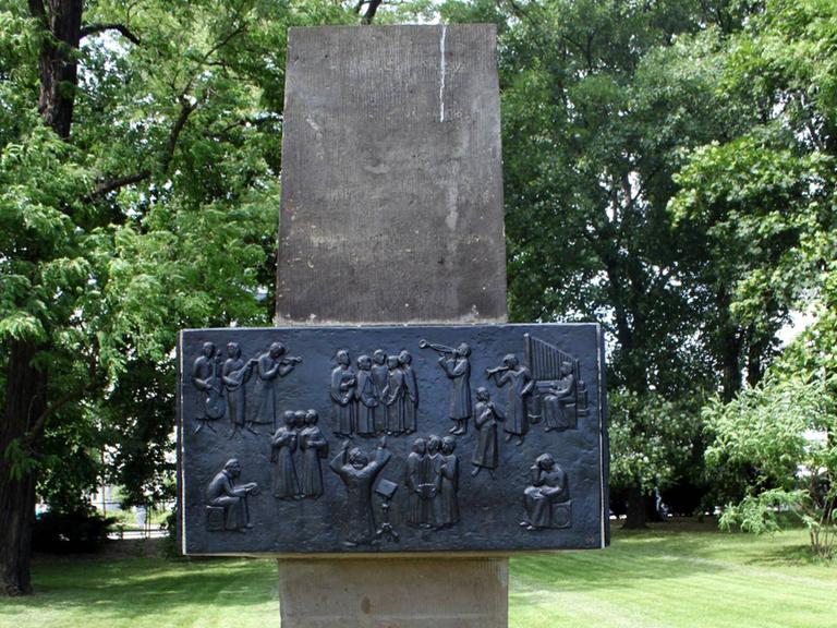 Auf dem Bild ist ein Denkmal aus einem großen senkrecht aufgestellten Stein zu sehen, auf dem eine schwarze Steintfafel montiert wurde. 