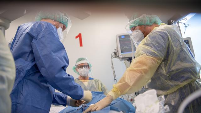 3 Ãrzte stehen vor einem Bett in einem Kranken-Haus. Sie tragen Schutz-Masken und spezielle Kleidung.