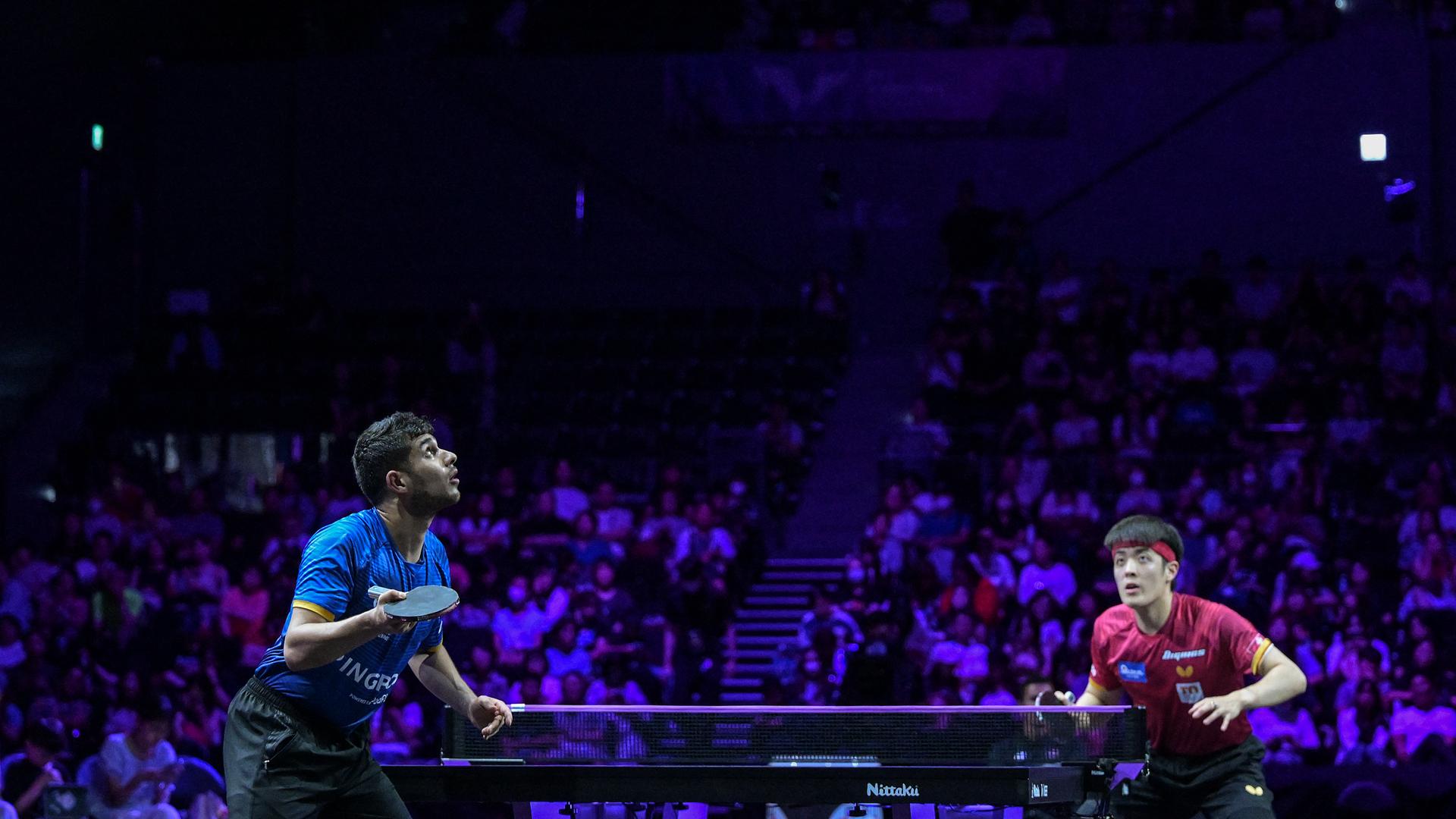 Zwei Spieler stehen um eine Tischtennisplatte in einer dunklen Sporthalle. Sie schauen nach oben zum Ball. Zwei Spieler stehen um eine Tischtennisplatte in einer dunklen Sporthalle. Sie schauen nach oben zum Ball.