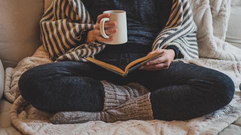 Eine Frau sitzt mit verschränkten Beinen in gemütlicher Kleidung auf einem Sofa und liest in einem Buch. In der Hand hält sie eine Tasse.