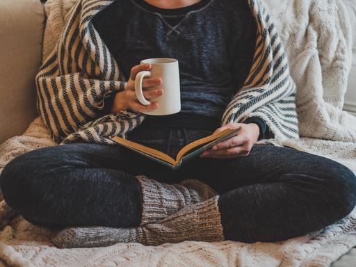 Eine Frau sitzt mit verschränkten Beinen in gemütlicher Kleidung auf einem Sofa und liest in einem Buch. In der Hand hält sie eine Tasse.
