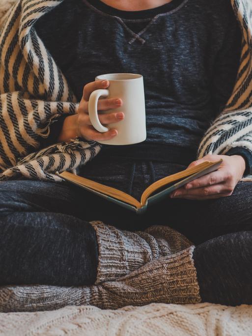 Eine Frau sitzt mit verschränkten Beinen in gemütlicher Kleidung auf einem Sofa und liest in einem Buch. In der Hand hält sie eine Tasse.