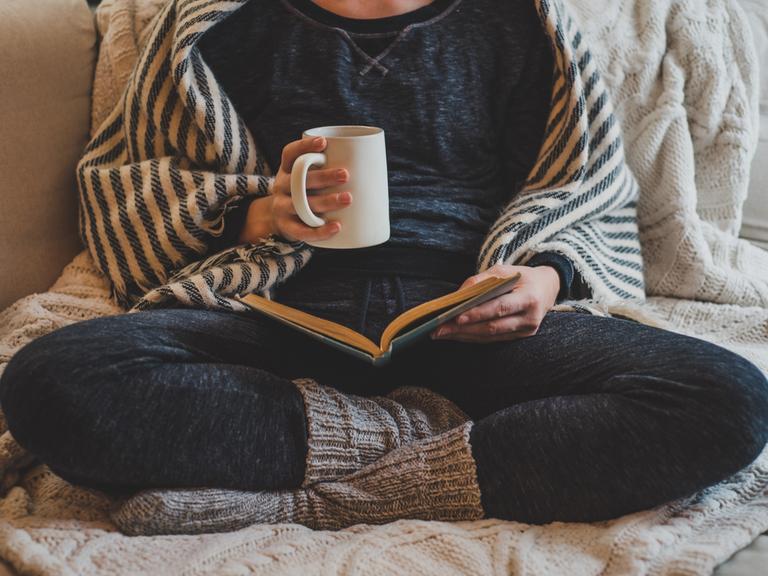 Eine Frau sitzt mit verschränkten Beinen in gemütlicher Kleidung auf einem Sofa und liest in einem Buch. In der Hand hält sie eine Tasse.