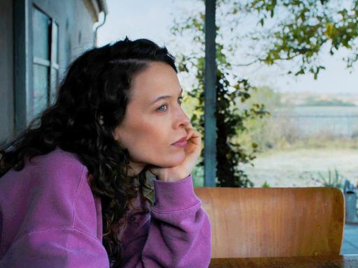 Paula Beer sitzt in pinkem Pullover an einem Tisch und schaut in die Ferne.