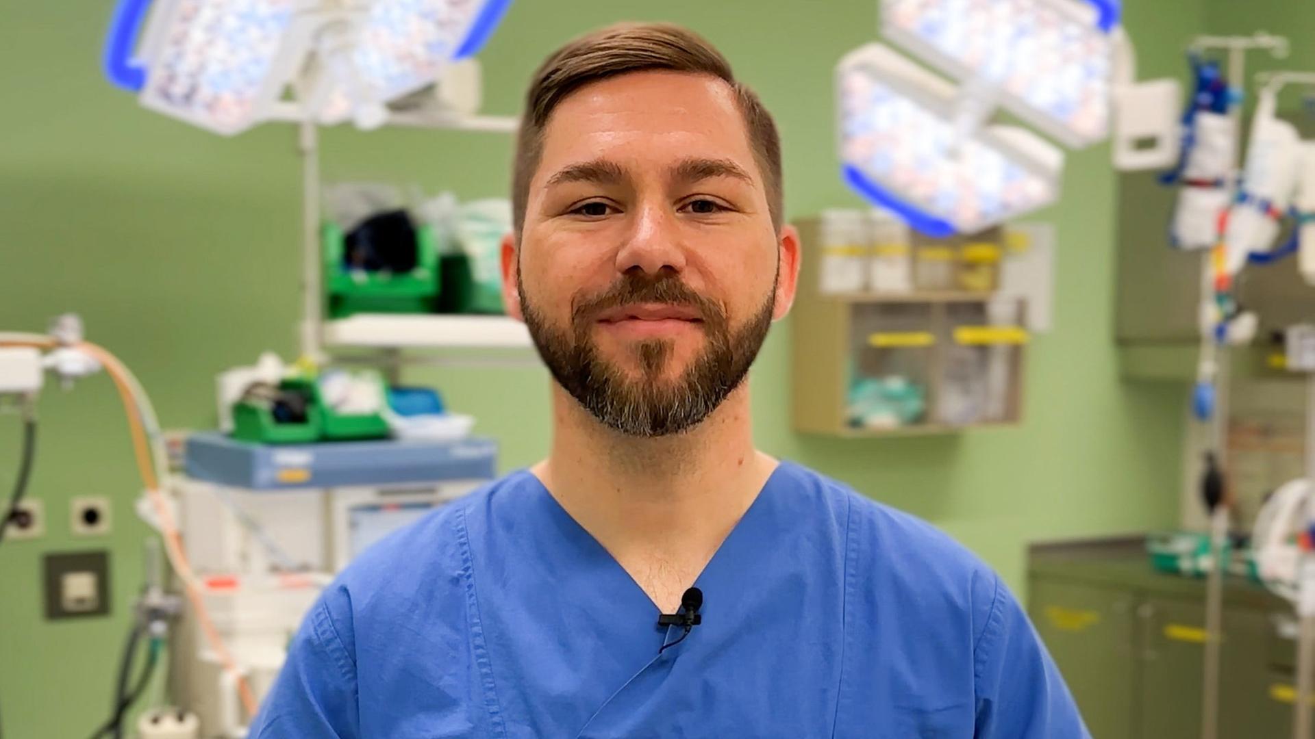 Dominik Stark steht in blauer Pflegerkleidung in eienm Krankenhauszimmer vor einer grünen Wand und einigen Geräten einer Intensivstation. Er schaut in die Kamera. Dominik Stark steht in blauer Pflegerkleidung in eienm Krankenhauszimmer vor einer grünen Wand und einigen Geräten einer Intensivstation. Er schaut in die Kamera.