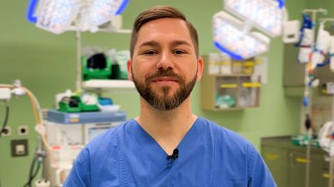 Dominik Stark steht in blauer Pflegerkleidung in eienm Krankenhauszimmer vor einer grünen Wand und einigen Geräten einer Intensivstation. Er schaut in die Kamera.