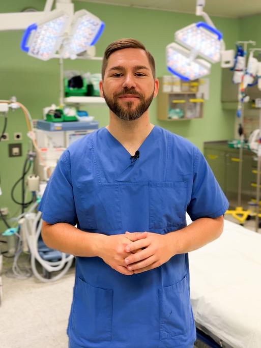 Dominik Stark steht in blauer Pflegerkleidung in eienm Krankenhauszimmer vor einer grünen Wand und einigen Geräten einer Intensivstation. Er schaut in die Kamera.