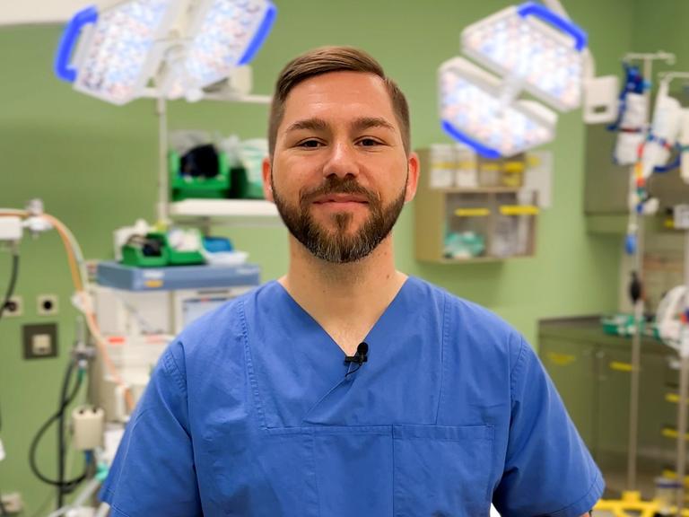 Dominik Stark steht in blauer Pflegerkleidung in eienm Krankenhauszimmer vor einer grünen Wand und einigen Geräten einer Intensivstation. Er schaut in die Kamera.