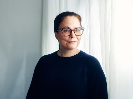Thea Dorn, mit Brille und dunklem Pullover, steht vor einem Fenster und blickt in die Kamera.