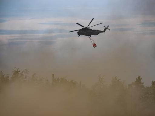 Ein Löschhubschrauber der Bundeswehr bei einem Waldbrand im Einsatz. An dem Hubschauber hängt ein Behälter für Löschwasser.