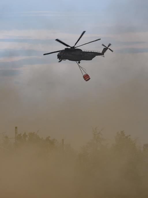 Ein Löschhubschrauber der Bundeswehr bei einem Waldbrand im Einsatz. An dem Hubschauber hängt ein Behälter für Löschwasser.