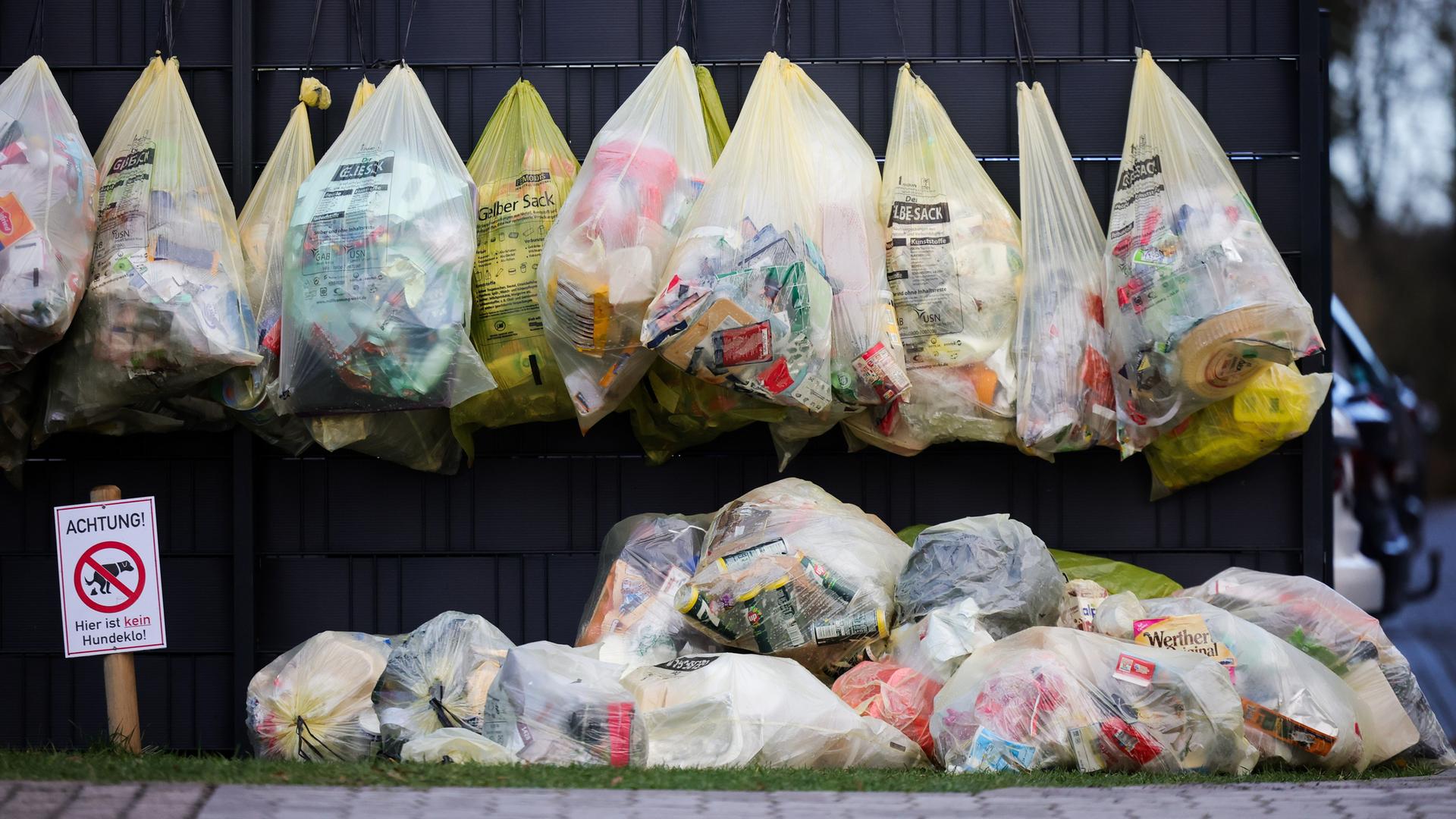 Gelbe Säcke mit Plastikmüll und Verpackungsmüll hängen an einem Zaun vor einem Mehrfamilienhaus.