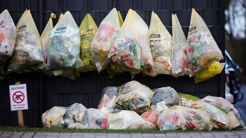 Gelbe Säcke mit Plastikmüll und Verpackungsmüll hängen an einem Zaun vor einem Mehrfamilienhaus.