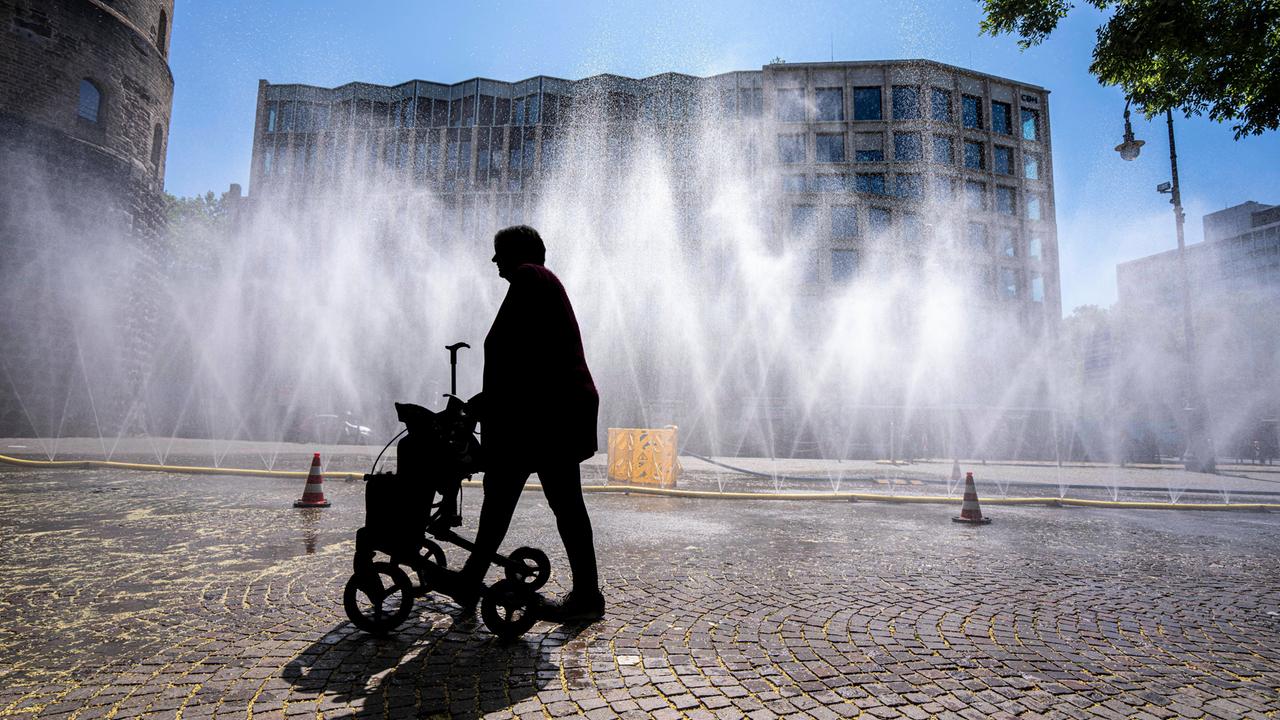 Eine Frau geht mit Rollator über den Rudolfplatz in Köln. Da hinter ihr Wasserschläuche für Abkühlung sorgen, ist sie nur als Silhouette zu erkennen.