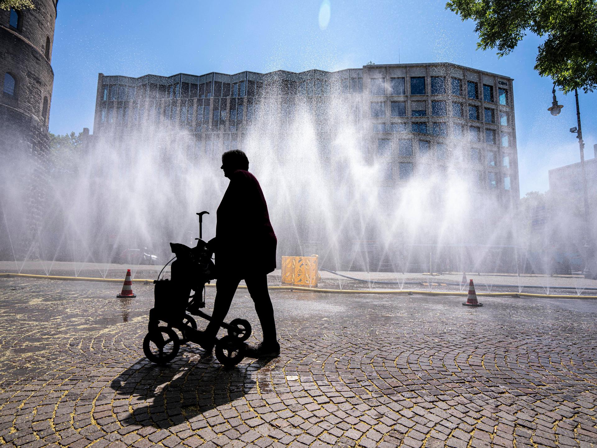 Eine Frau geht mit Rollator über den Rudolfplatz in Köln. Da hinter ihr Wasserschläuche für Abkühlung sorgen, ist sie nur als Silhouette zu erkennen.