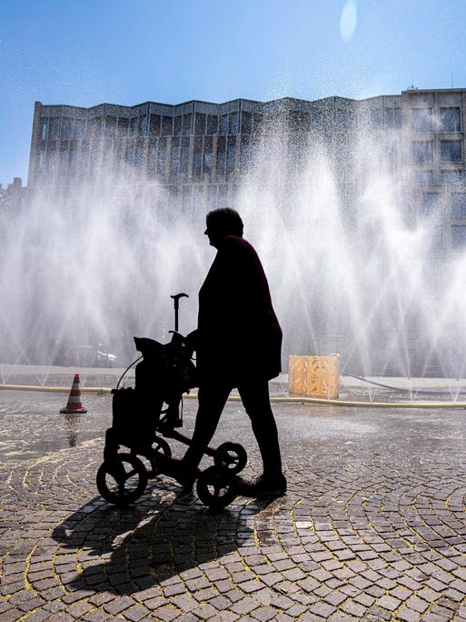 Eine Frau geht mit Rollator über den Rudolfplatz in Köln. Da hinter ihr Wasserschläuche für Abkühlung sorgen, ist sie nur als Silhouette zu erkennen. Eine Frau geht mit Rollator über den Rudolfplatz in Köln. Da hinter ihr Wasserschläuche für Abkühlung sorgen, ist sie nur als Silhouette zu erkennen.