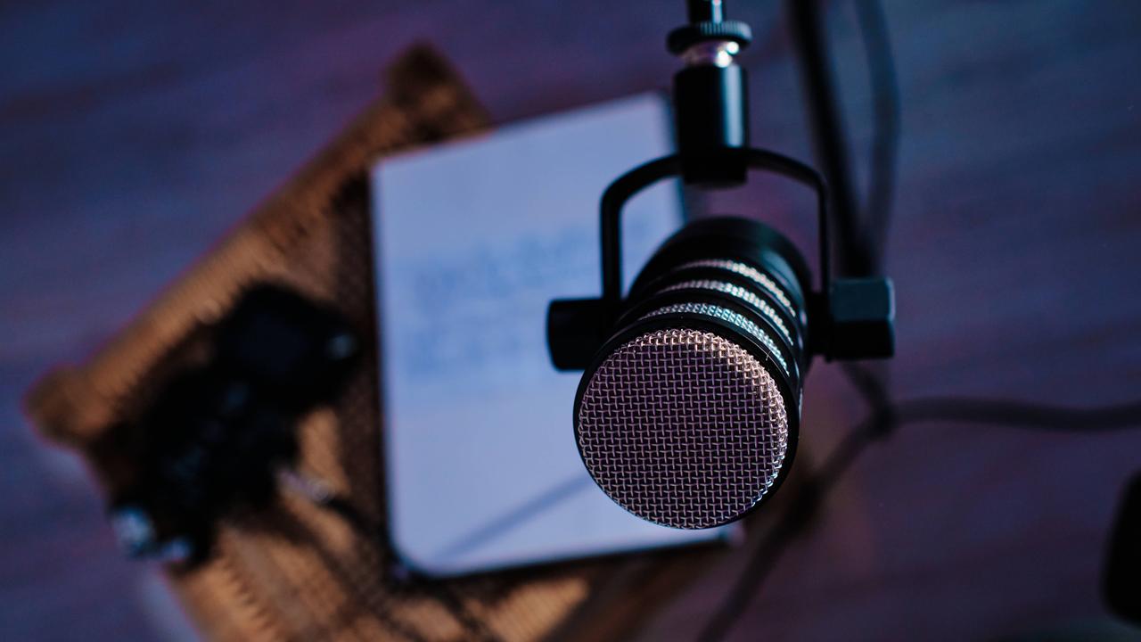 Das Bild zeigt eine Detailaufnahme eines schwarzen Mikrofons. Es hängt in einer Halterung. Im Hintergrund ist ein Bildschirm zu erkennen.